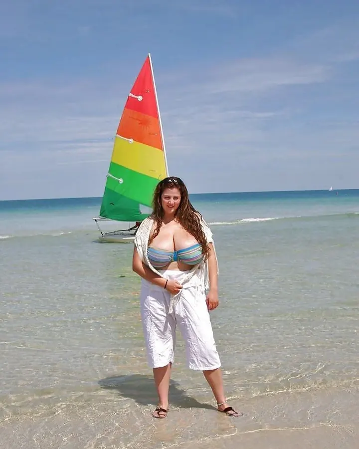 Femme aux cheveux châtains, foulard clair et maillot à rayures, debout dans l'eau devant une voile colorée.