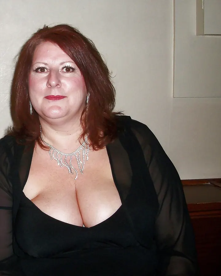 Femme rousse en robe noire décolletée, collier argenté, fond clair.