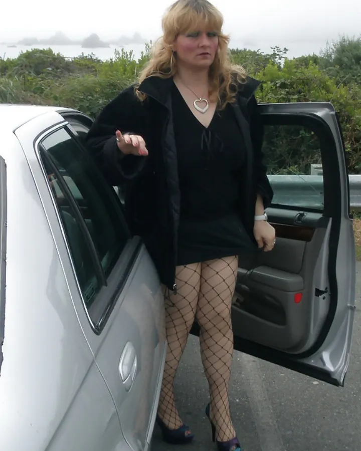 Femme blonde sortant d'une voiture, collants résille, veste noire, bord de mer brumeux.