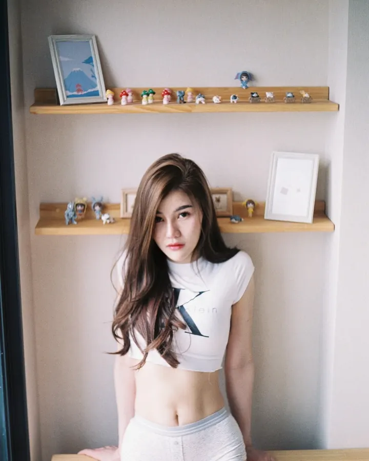Femme asiatique en t-shirt blanc court et short clair, mains posées sur un meuble, étagères en arrière-plan.