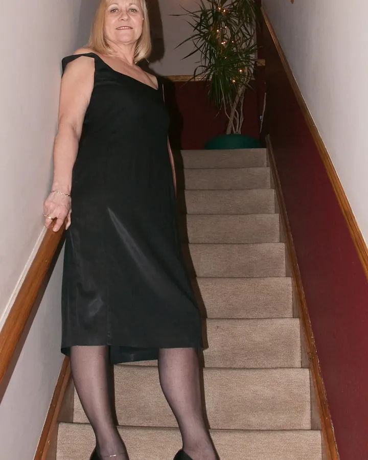 Femme blonde en robe noire assise dans un escalier, jambes croisées, sourire discret