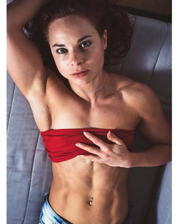 Femme brune aux yeux clairs, chemise noire et veste rose pâle, assise sur des marches dehors.