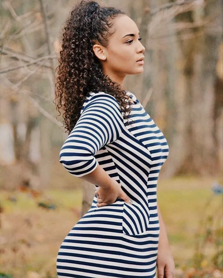Femme de profil en robe rayée bleu et blanc, cheveux bouclés relevés, mains sur les hanches