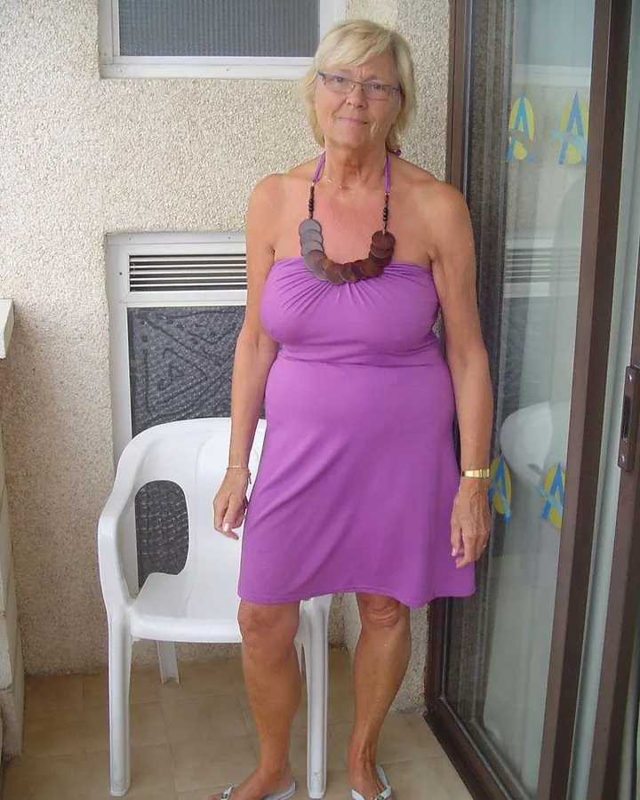 Femme aux cheveux platine, lunettes, robe violette, debout sur une terrasse près d'une chaise blanche