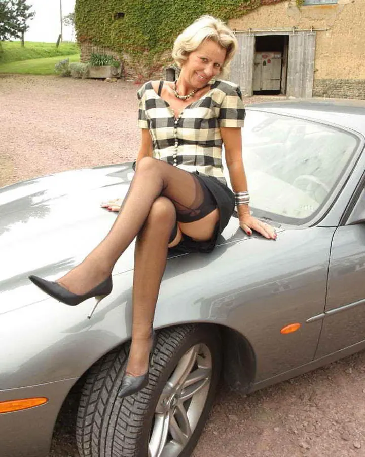 Femme aux cheveux blonds assise sur le capot d'une voiture grise, tailleur à carreaux, bas résille.