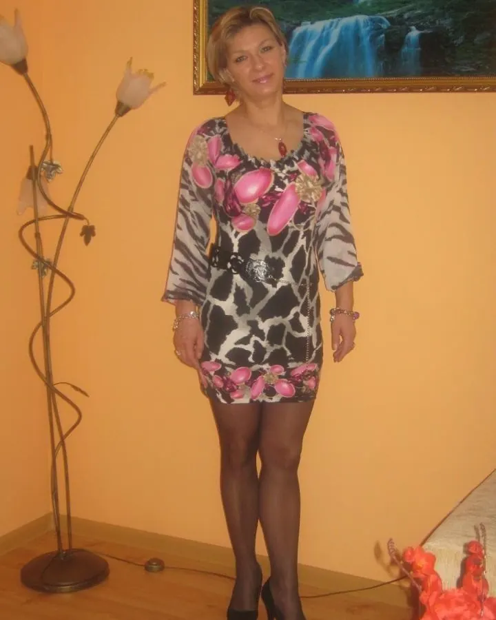 Femme blonde en robe imprimée, debout dans un salon aux murs orangés.