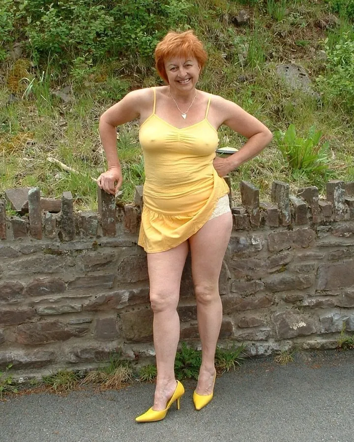 Femme aux cheveux roux en robe jaune, appuyée contre un muret de pierre.