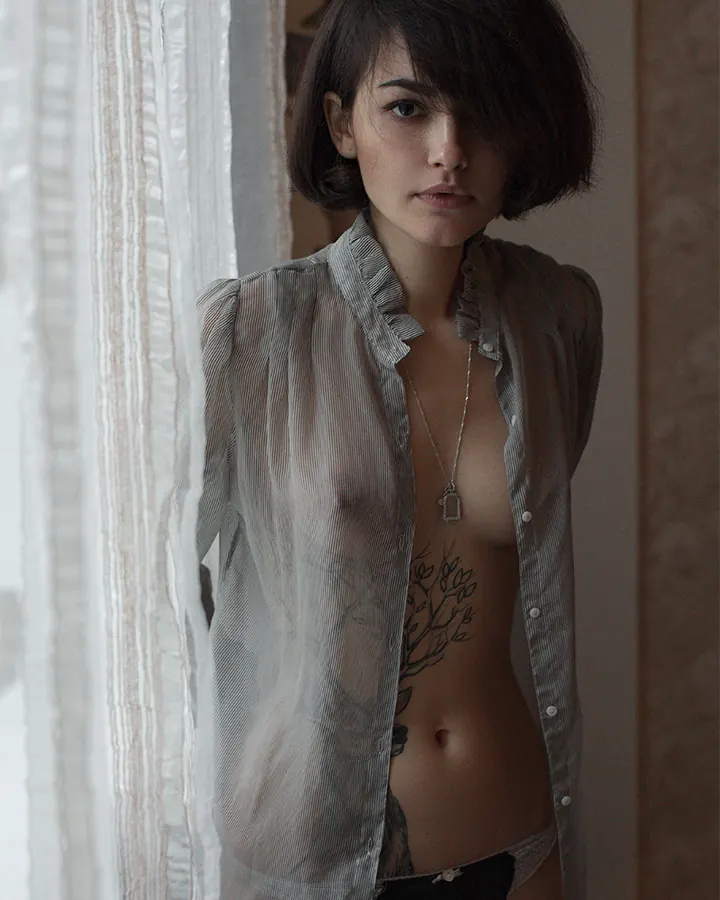Femme brune aux yeux bleus, chemise ouverte, debout près d'un rideau clair.