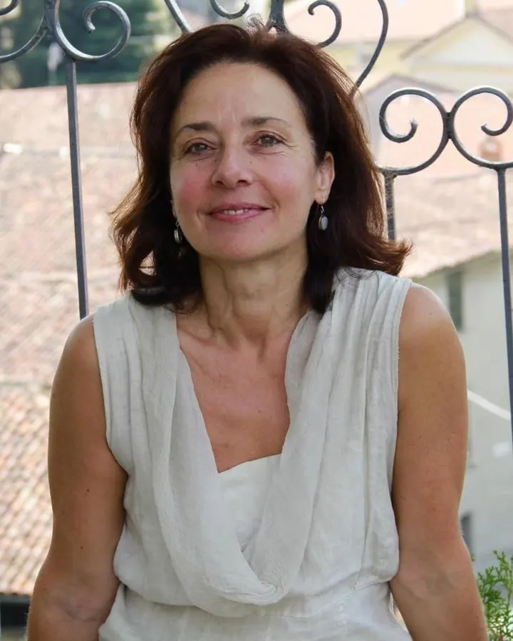 Femme aux cheveux châtains, chemise claire, sourire léger devant un mur clair.