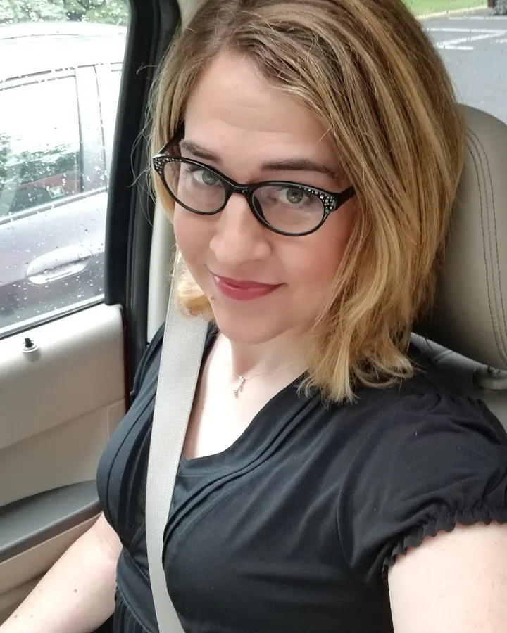 Femme blonde assise dans un canapé, souriant, lunettes fines et robe noire.