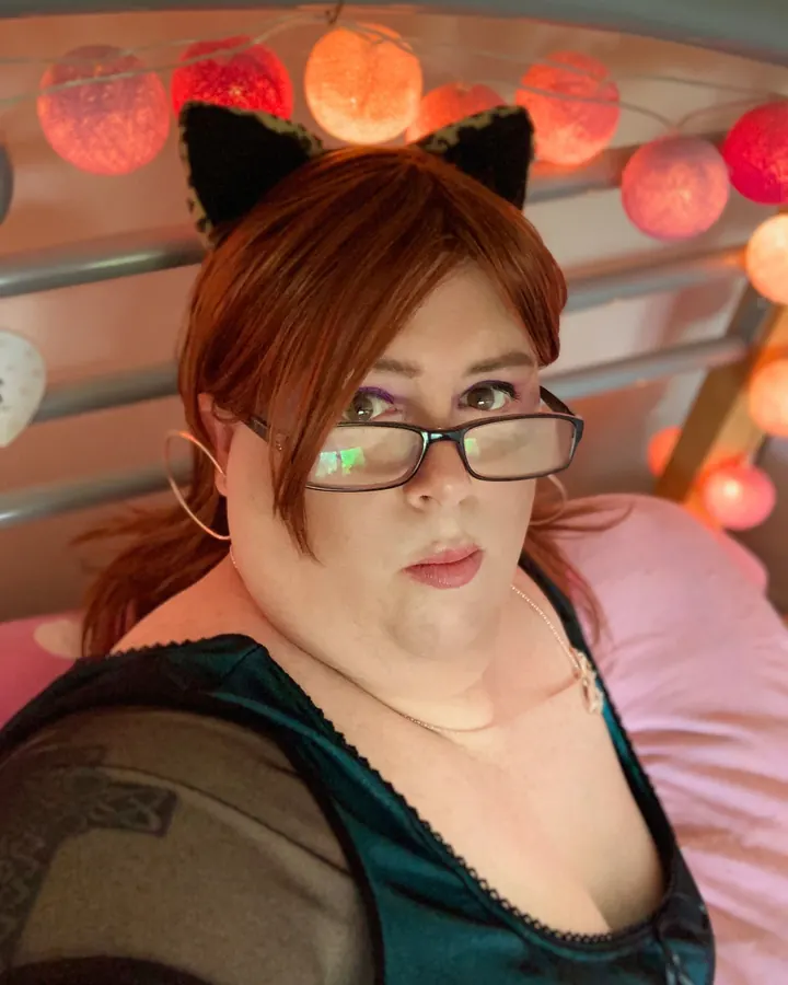 Femme ronde aux cheveux roux avec lunettes et serre-tête noir, assise sur un lit, guirlande lumineuse