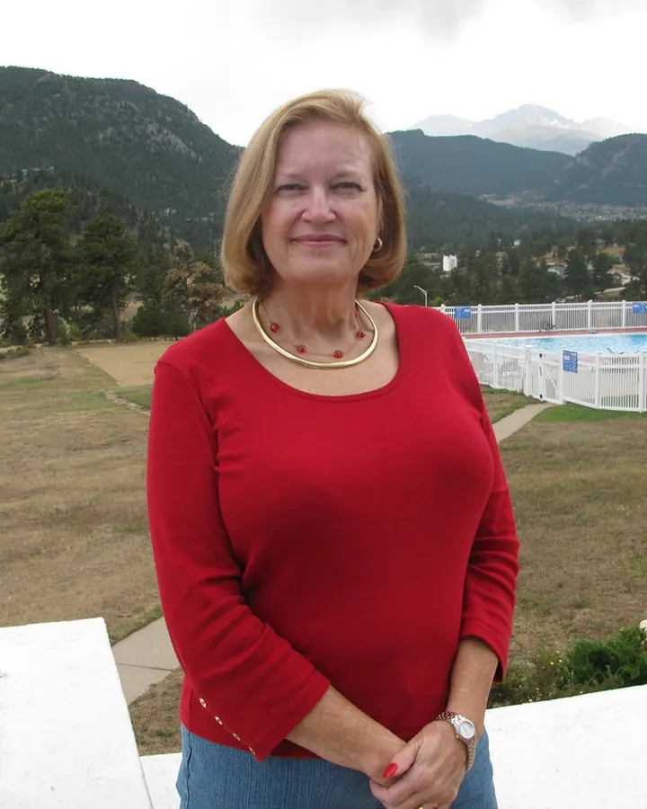 Femme aux cheveux blonds courts, pull rouge, debout devant un paysage de montagnes et une piscine