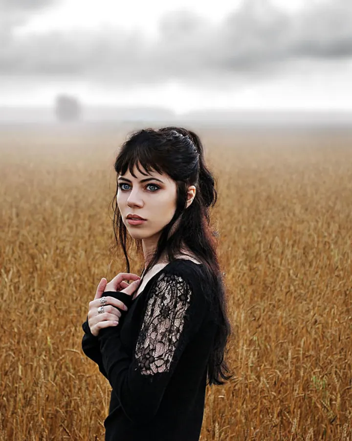 Femme en noir dans un champ de blé sous un ciel gris, cheveux noirs, regard direct