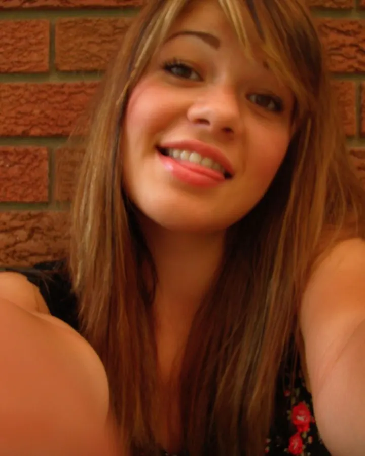 Jeune femme aux cheveux châtains devant un mur de briques rouges, large sourire, bras tendus.